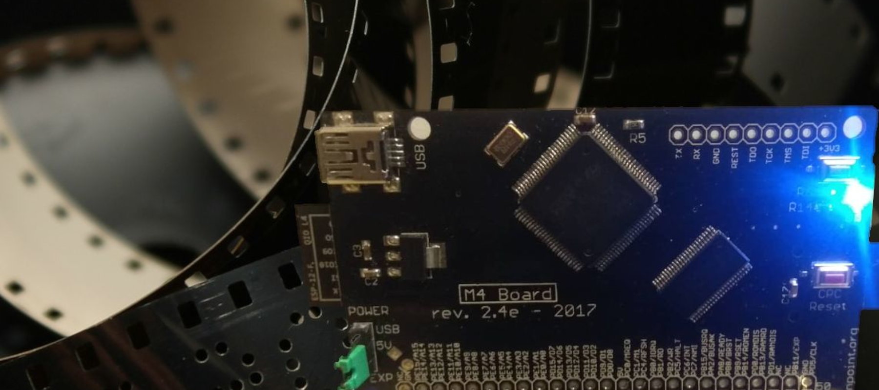 Close-up of a board labeled 'M4 Board Rev. 2.4e - 2017' and electronic components attached. A blue diode is visible on the right side of the board. The background is blurred, resembling a film strip.