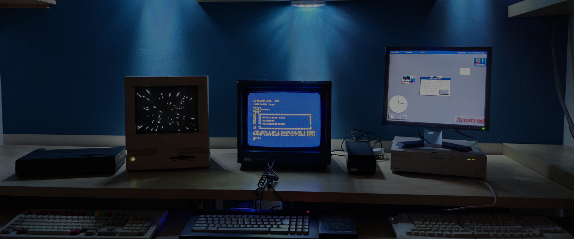 The office has a desk and three old desktop computers and monitors on it with keyboards on the shelf under the desk. The background features a blue wall with spotlights from above.