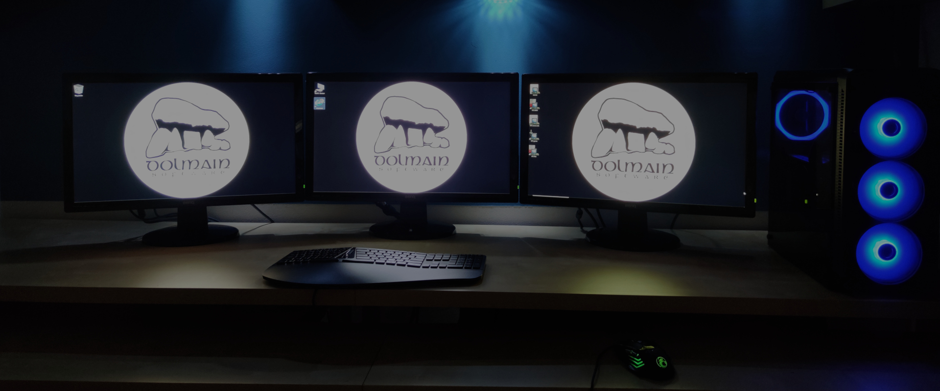 Modern home office with three computer monitors arranged in a triangle on a wooden desk. Each monitor displays a white logo in a circle, with the text 'Dolmain Software' in the center. A black keyboard and mouse sit in front of the desk, and a computer tower with three blue lights sits to the right. In the background is a blue wall with moody spotlights from above