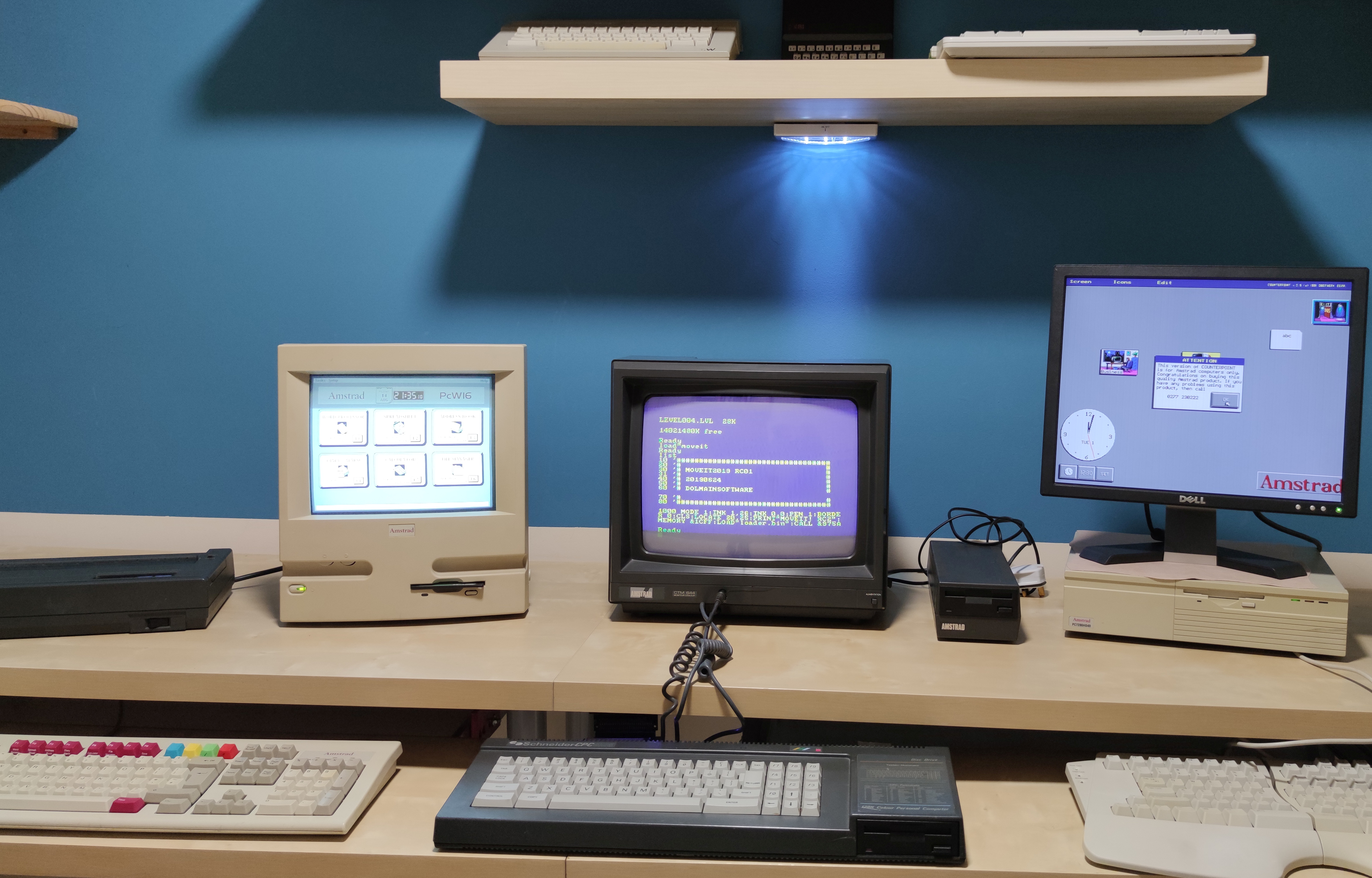 The office has a desk and three old desktop computers and monitors on it with keyboards on the shelf under the desk. The background features a blue wall with spotlights from above.